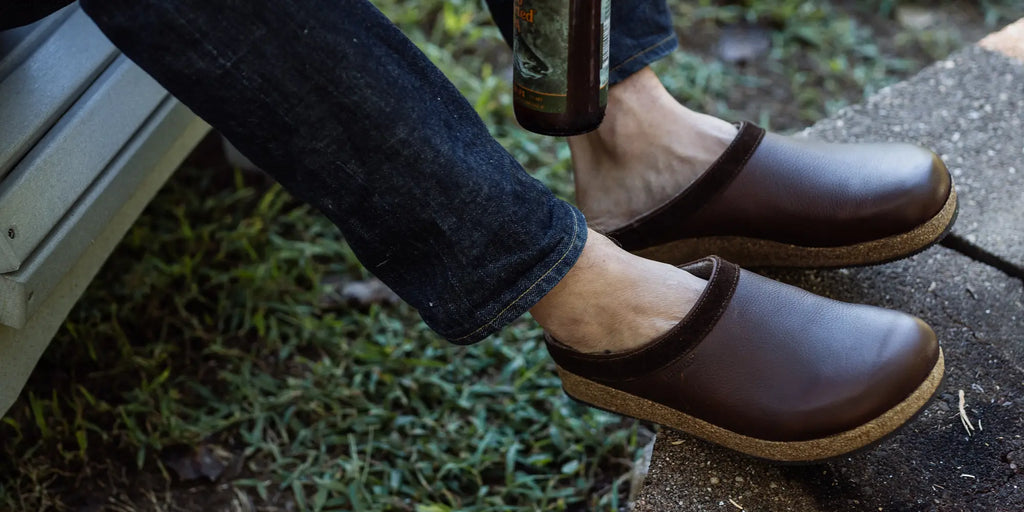 A person wearing brown slip-on shoes with cork soles sits on a bench outdoors, their jeans rolled up, holding a glass bottle by their side.