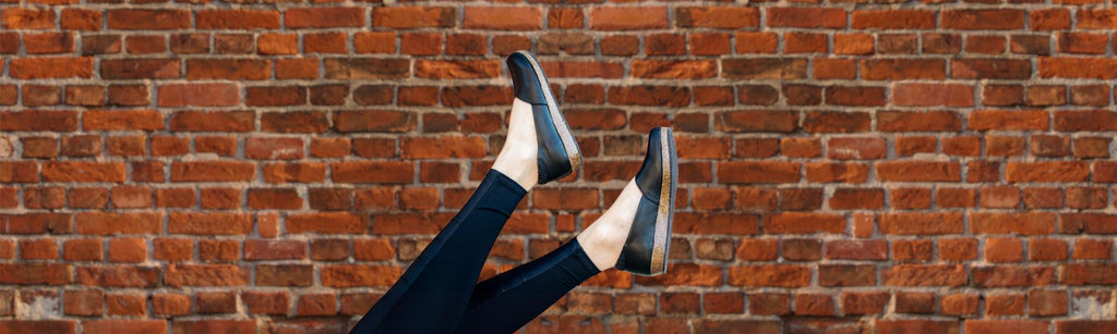 Feet wearing black shoes and black pants are playfully raised against a red brick wall background.