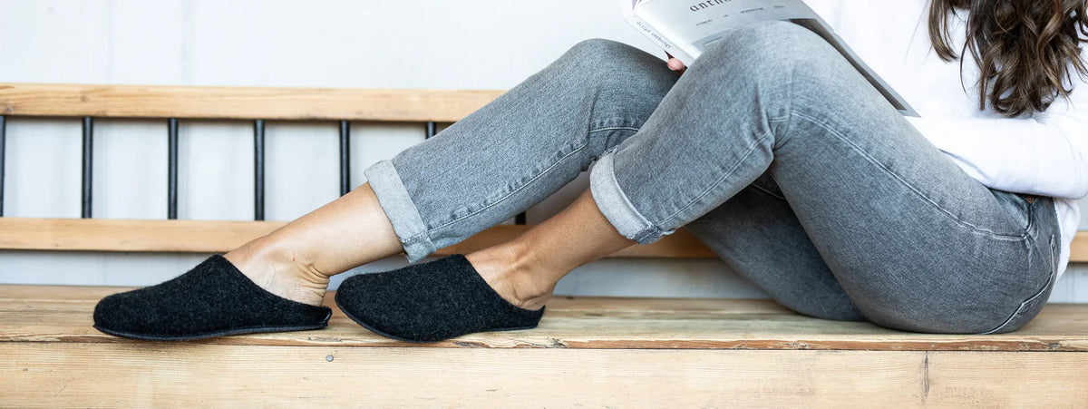 Person sitting on a wooden bench wearing gray jeans and black slippers, holding an open book, with legs stretched out and slightly crossed.