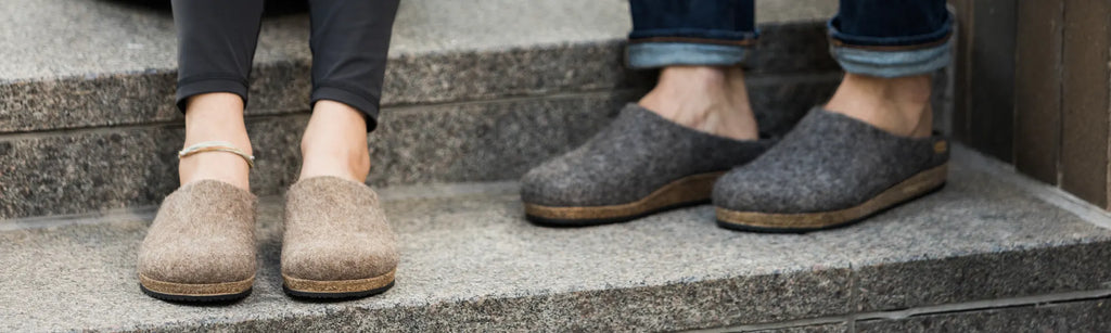 Two people wearing wool slippers, one in green and one in red, relax on a bed.