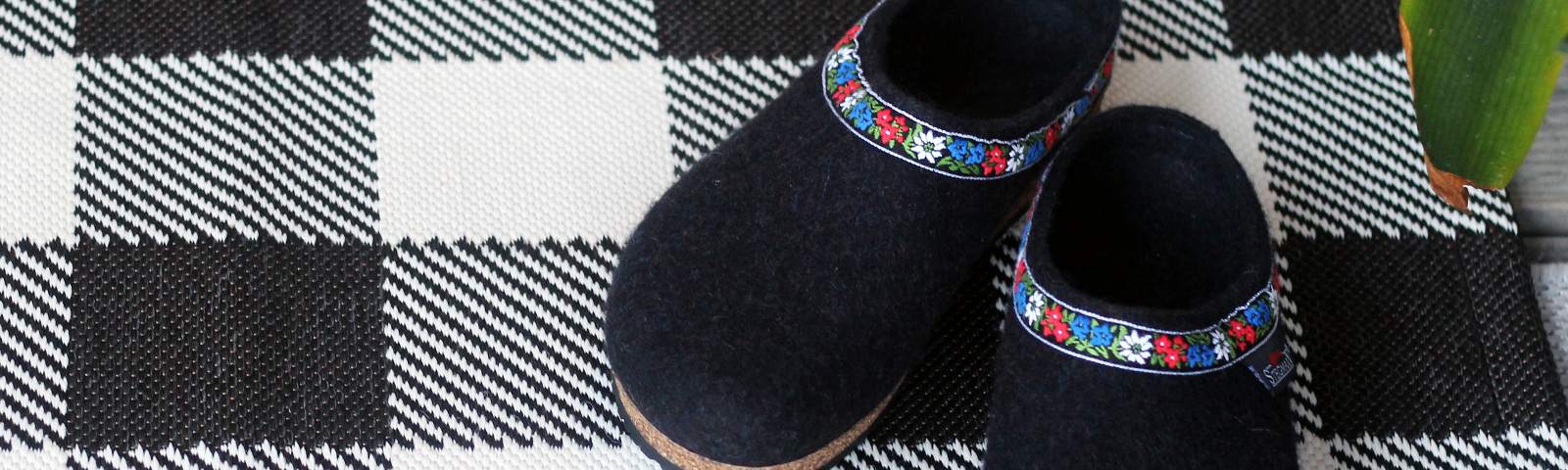 A pair of black felt slippers with floral trim on a black and white checkered mat, with a green plant partially visible in the corner.