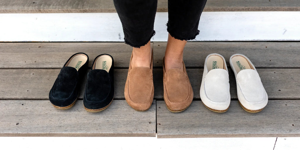 Person wearing brown suede slip-on shoes stands on wooden steps, with a pair of black shoes on the left and a pair of white shoes on the right.