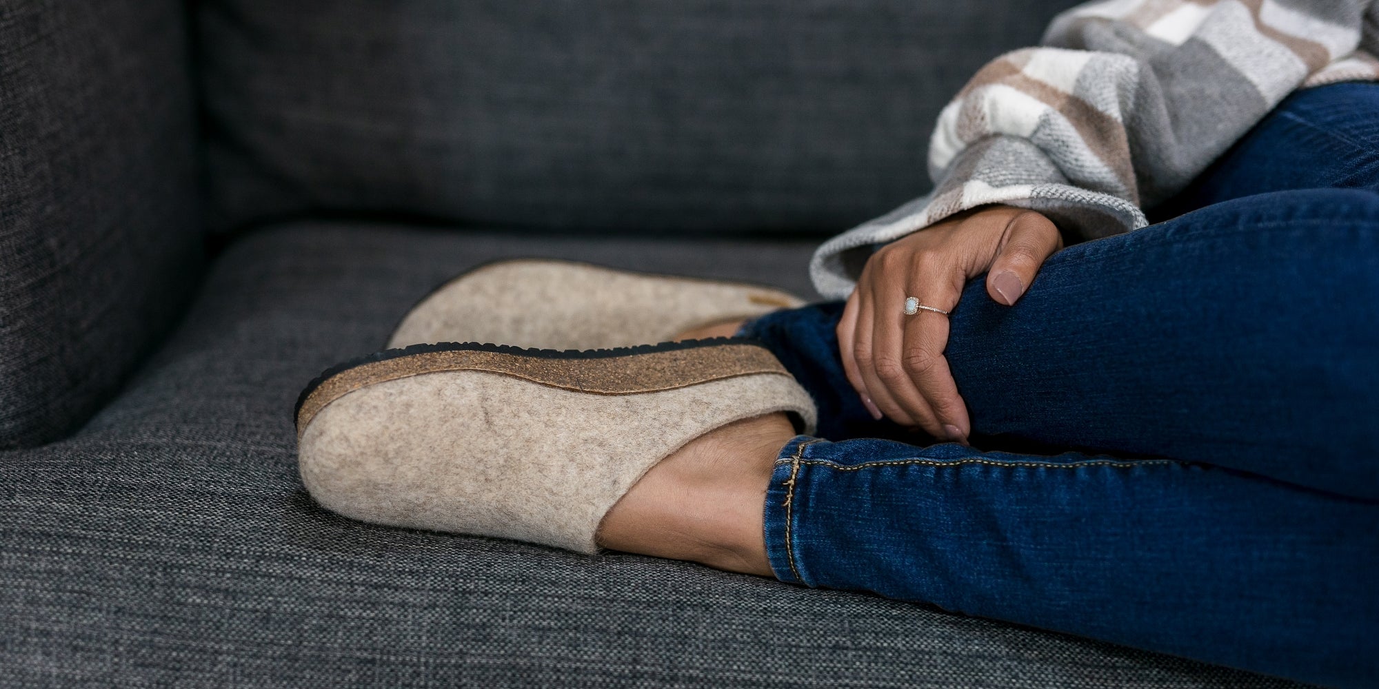 Person wearing gray wool slippers with cork soles and dark jeans, sitting near stacked firewood on a light-colored floor.