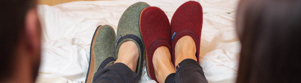 Two people wearing wool slippers, one in green and one in red, relax on a bed.