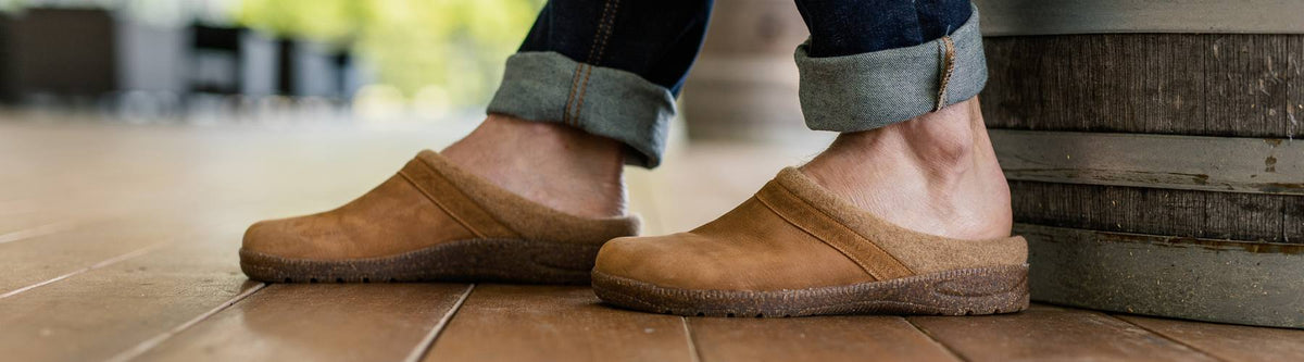 A person wearing rolled-up jeans and brown slip-on clogs stands on a wooden floor next to a large barrel.