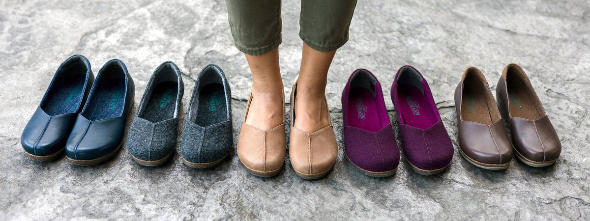 A person stands on a stone surface wearing tan slip-on shoes, with additional pairs of similar shoes in various colors arranged on either side.