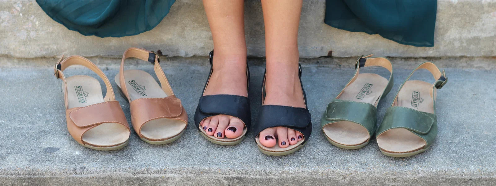 A person stands on a stone surface wearing tan slip-on shoes, with additional pairs of similar shoes in various colors arranged on either side.