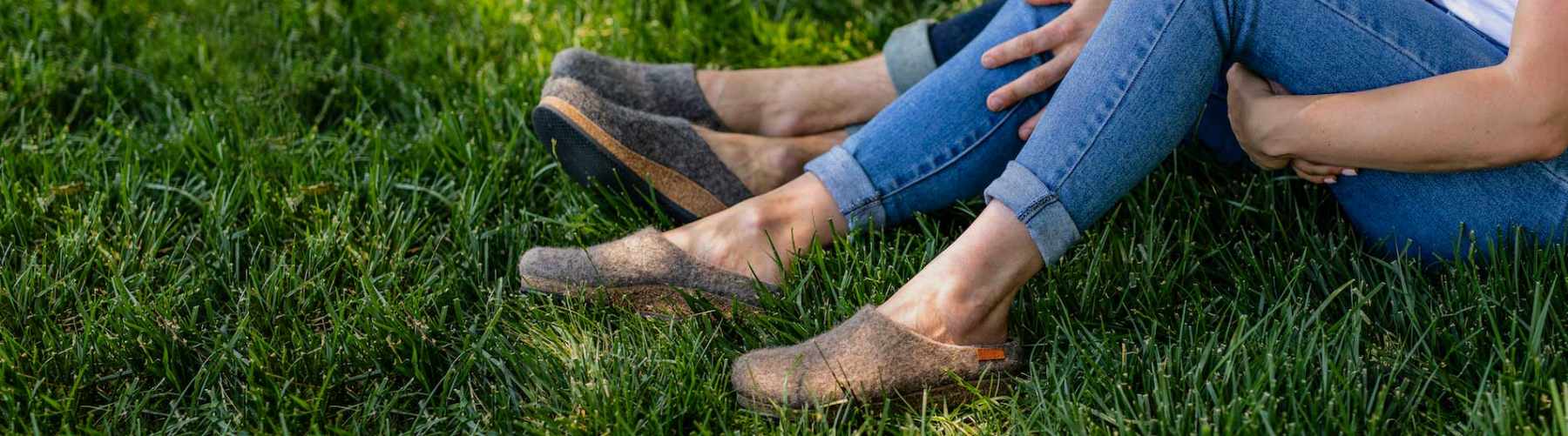 Two people sitting on grass wearing gray slip-on shoes and blue jeans, with only their legs and feet visible.