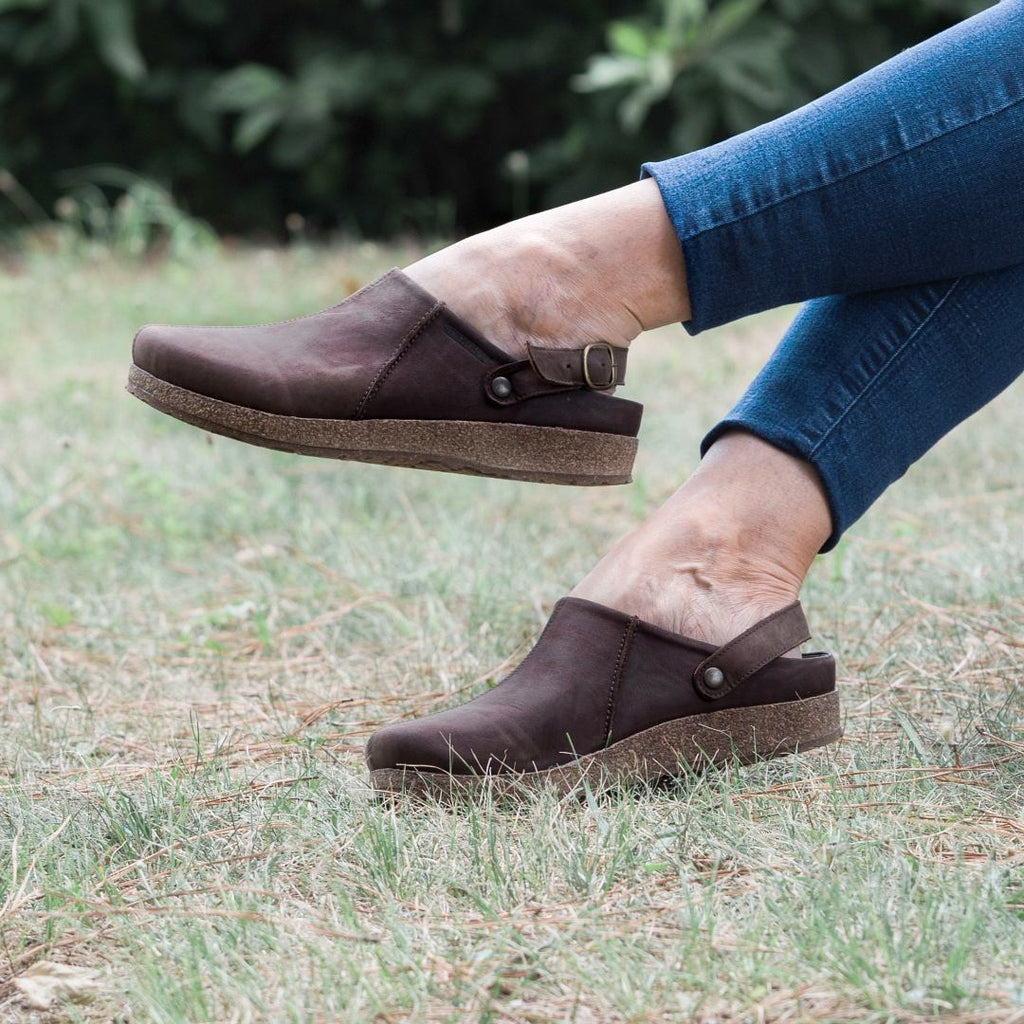 A person in casual jeans sits on the grass, wearing Women's Tirol Leather Clog in brown, made from oiled leather.
