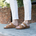 A person in white pants and tan Women's Heidi Leather Sandals with adjustable straps stands on concrete before a brick wall and green foliage.