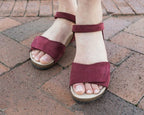A person stands on a brick pavement wearing Women's Freya Leather Wedge Sandals—red, open-toe with ankle straps and arch support.