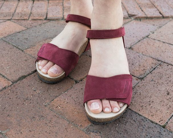 A person stands on a brick pavement wearing Women's Freya Leather Wedge Sandals—red, open-toe with ankle straps and arch support.