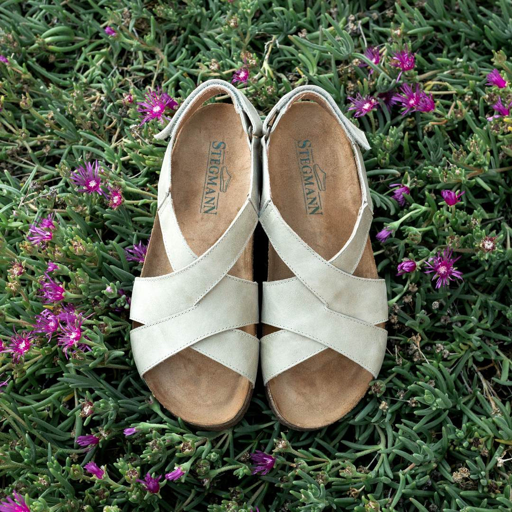 A pair of Women's Hannah Leather Sandal - Wide in light beige, featuring a cushioned footbed and sustainable cork midsole, placed side by side on green foliage with small purple flowers.