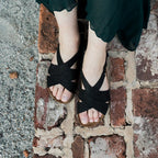 Standing on a rustic brick surface, a person wears the elegant Women's Hannah Leather Sandal - Medium, featuring a black strappy design and cushioned suede footbed. A flowing dark green skirt complements this chic footwear.