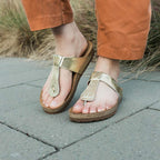 Feet wearing Women's Inga Leather Sandals on a stone path, paired with brown pants. The signature sustainable cork footbed offers comfort and style, with lush grass visible in the background.