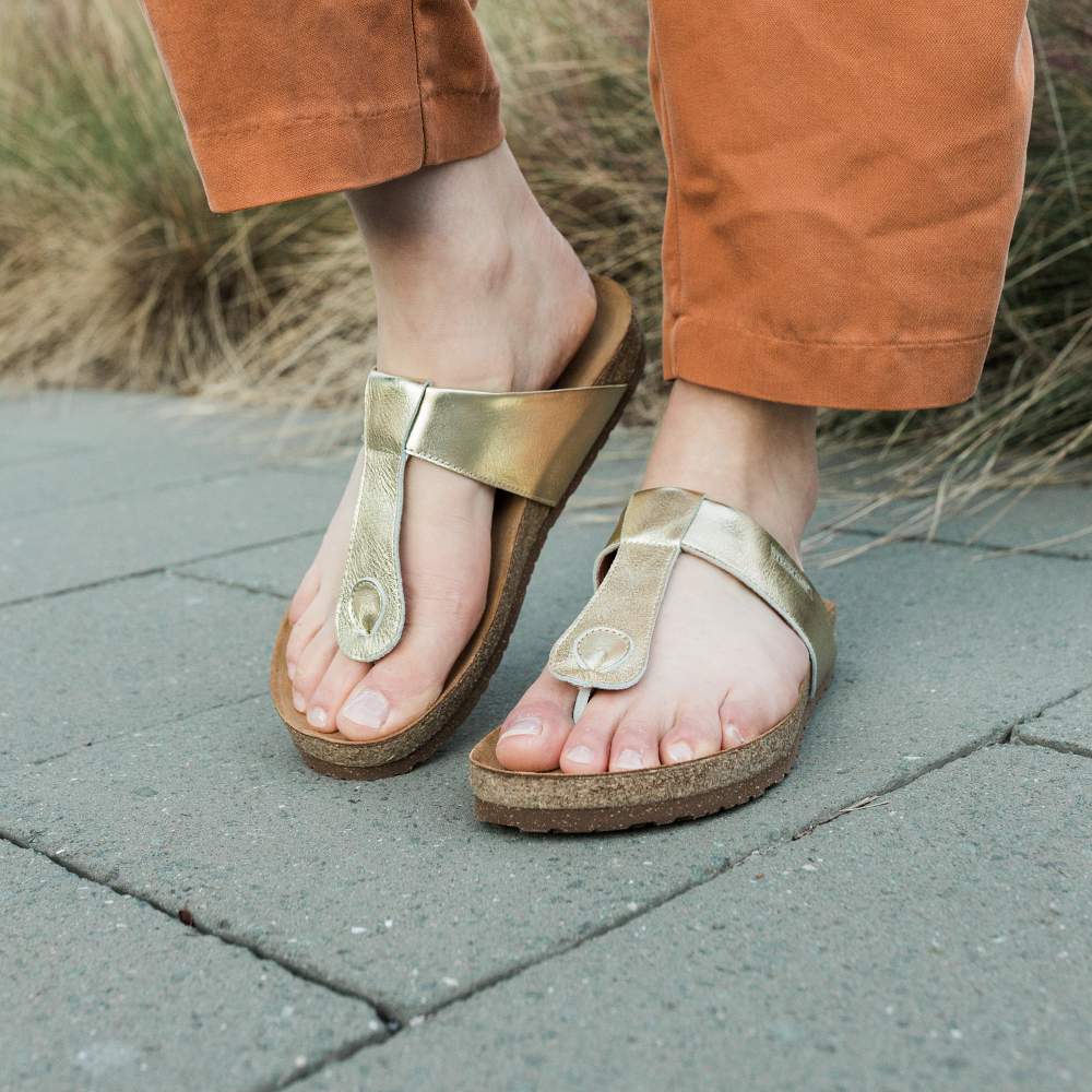 Feet wearing Women's Inga Leather Sandals on a stone path, paired with brown pants. The signature sustainable cork footbed offers comfort and style, with lush grass visible in the background.