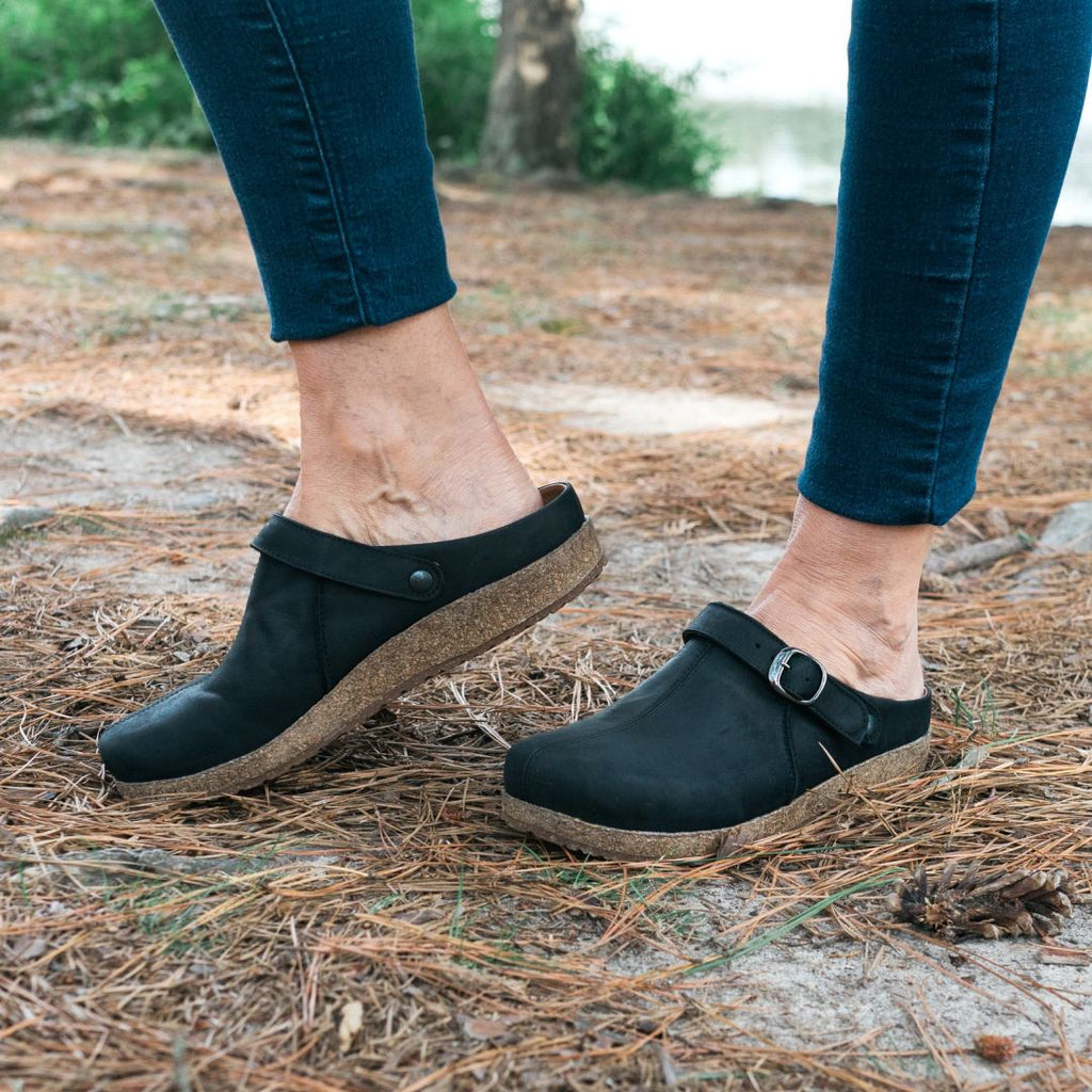 A person stands outdoors on a pine needle-covered ground, showcasing Women's Tirol Leather Clog with adjustable straps and cork soles. The design features a wide toe box, blending comfort and style for any outdoor adventure.