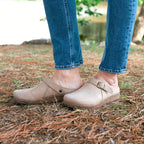 Standing on pine needle-covered ground, a person showcases light brown Women's Tirol Leather Clog slip-on shoes. Crafted from oiled leather and paired with jeans, these clogs offer a wide toe box for enhanced comfort.