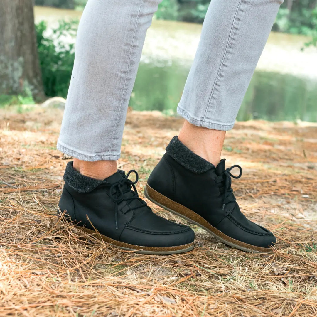 A person wearing Women's Baden Leather Boot in black oiled leather with ultra cushion support insoles and light gray jeans stands on pine needles outdoors.