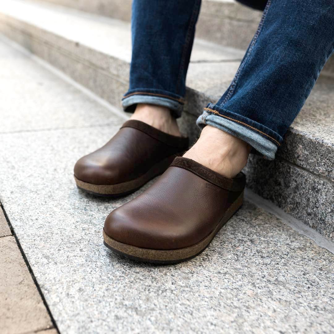 A person wearing Men's Graz Leather Clog with a cork footbed and rolled-up jeans sits on concrete steps.