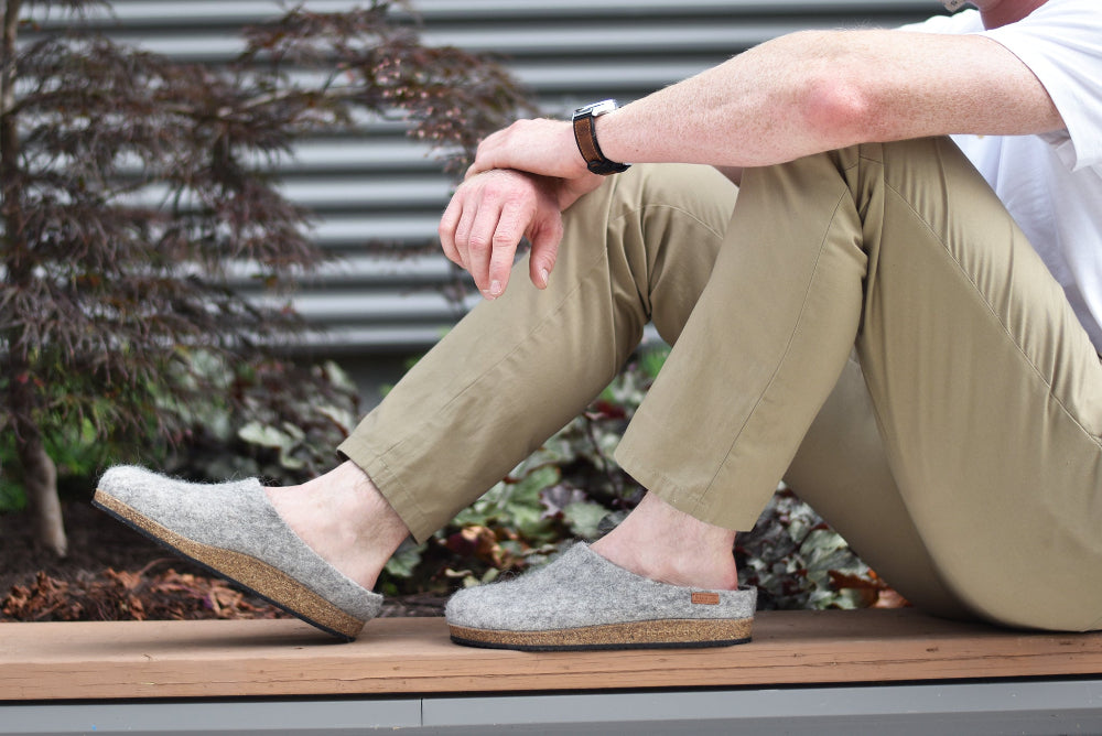 A person wearing tan pants, a white shirt, and Men's Eco Wool Clog slippers sits on a wooden bench outdoors near plants.