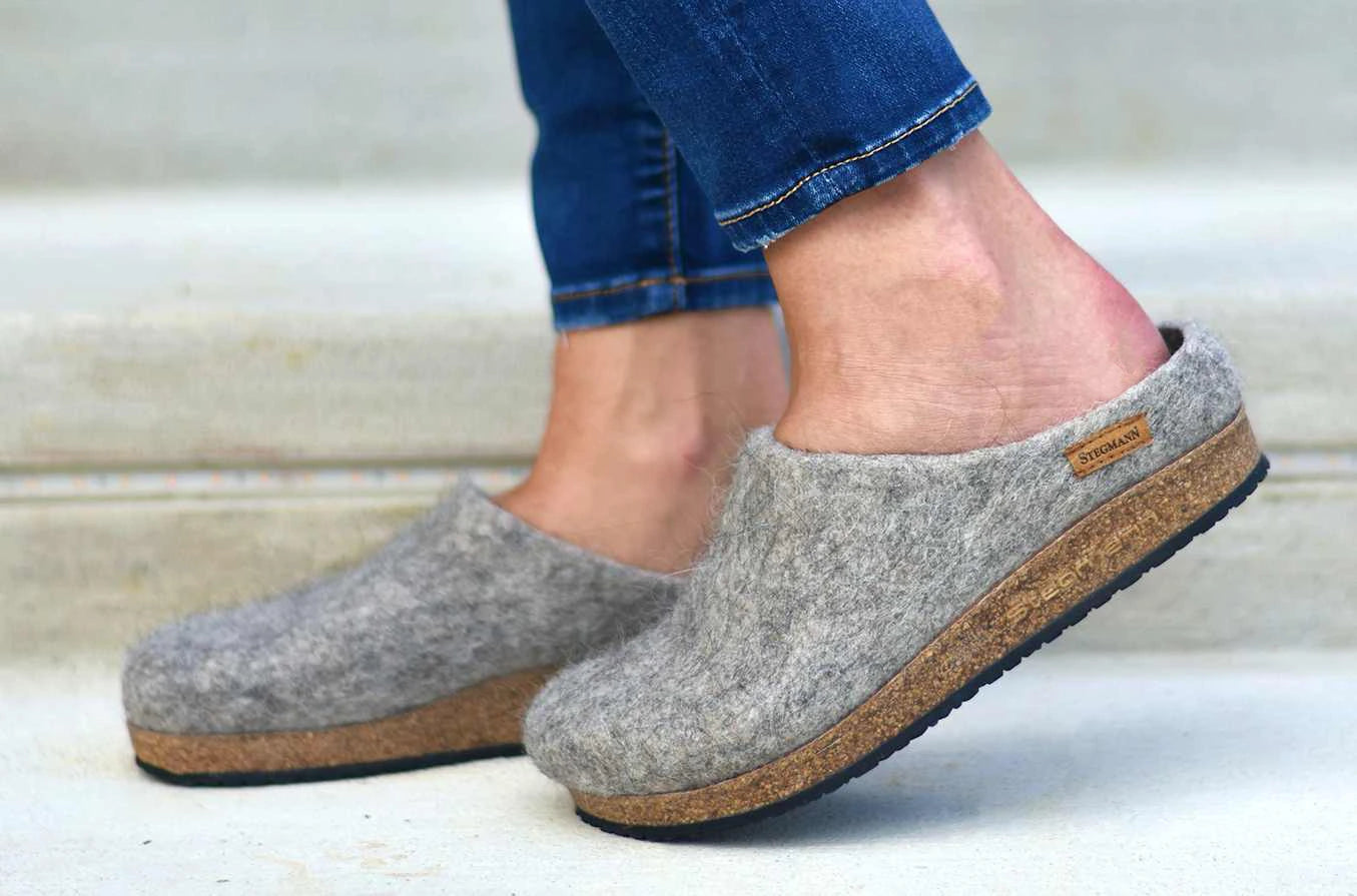 A person wearing Women's Eco Wool Clog slippers with a cork latex footbed and blue jeans stands on light steps—an effortless showcase of sustainable wool footwear.