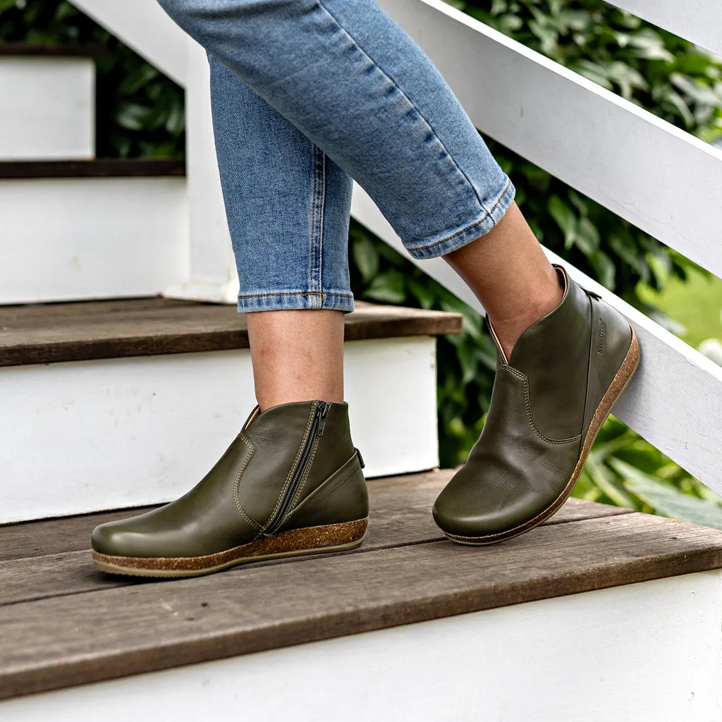 A woman wearing the Women's Leather Amalia Boot—green ankle boots with zippers and light brown soles—stands on outdoor wooden steps in blue rolled-up jeans.