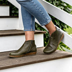 A woman wearing the Women's Leather Amalia Boot—green ankle boots with zippers and light brown soles—stands on outdoor wooden steps in blue rolled-up jeans.