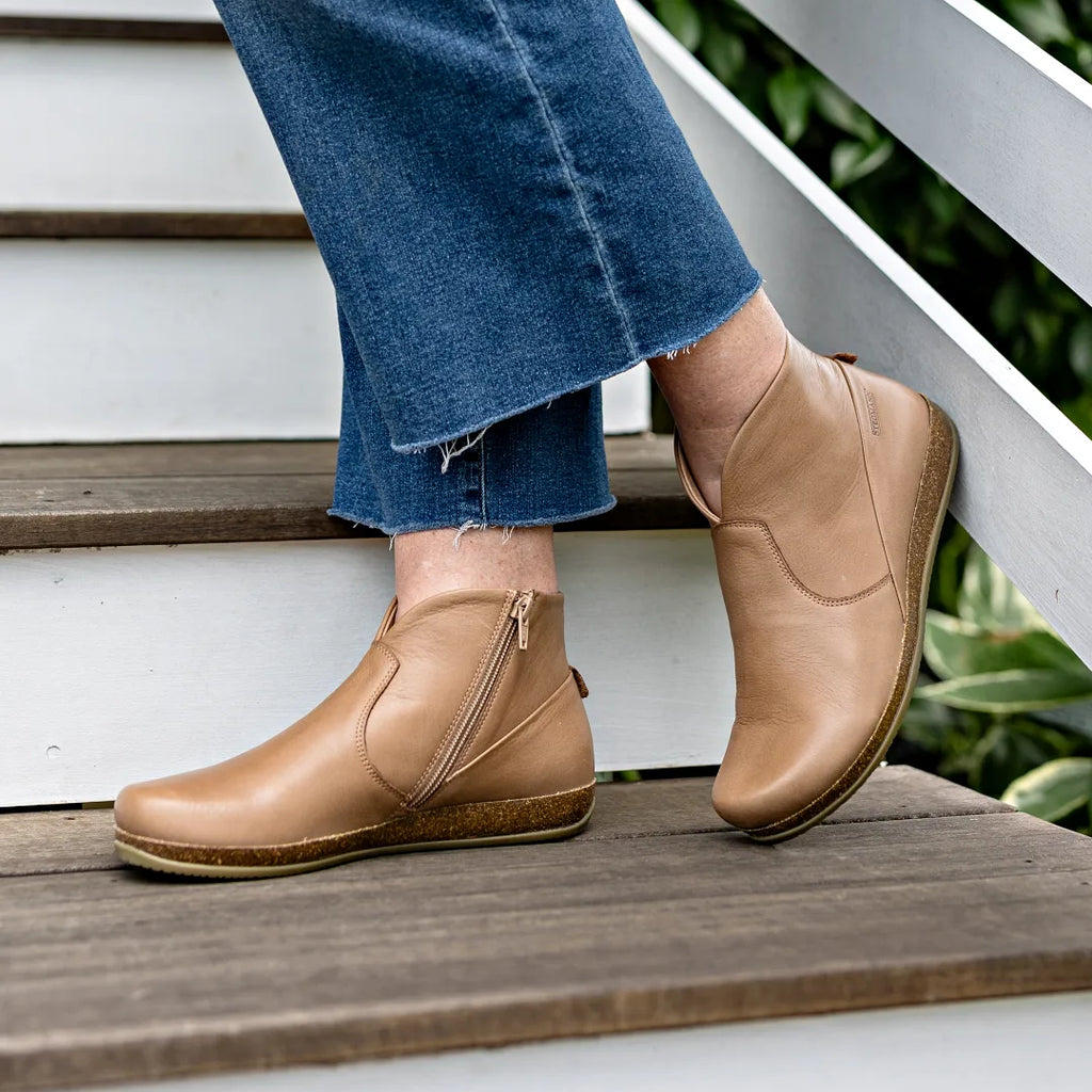 A person wearing the Women's Leather Amalia Boot in tan with side zippers and blue jeans stands on white wooden steps.