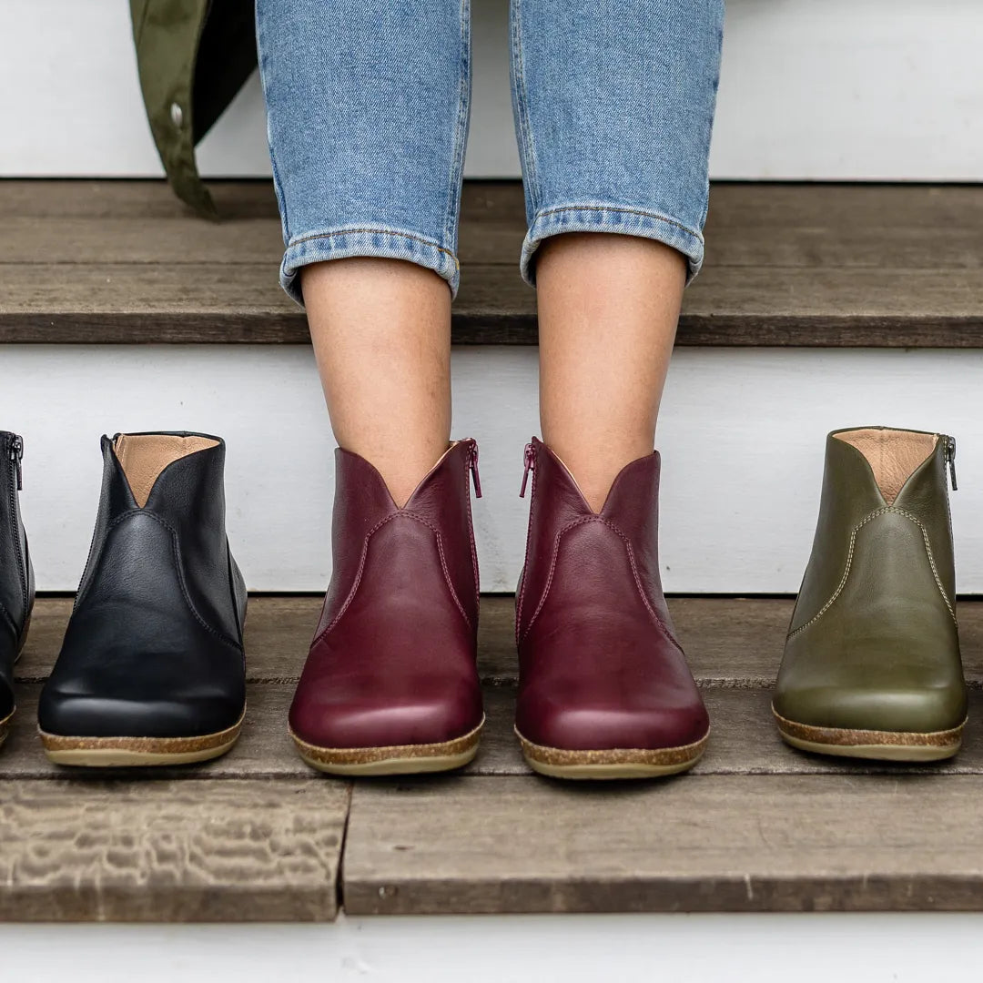 A person in blue jeans and maroon Women's Leather Amalia Boots stands on wooden steps, with black and olive green Women's Leather Amalia Boots placed on either side.