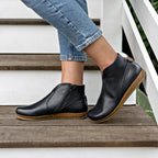A person wearing cropped blue jeans and Women's Leather Amalia Boot stands on wooden stairs next to a white railing.