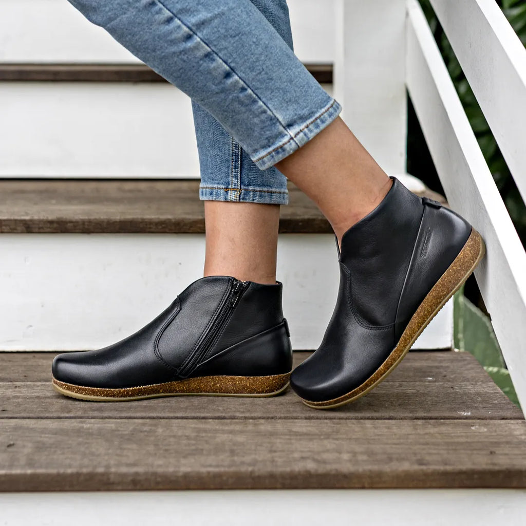 A person wearing cropped blue jeans and Women's Leather Amalia Boot stands on wooden stairs next to a white railing.
