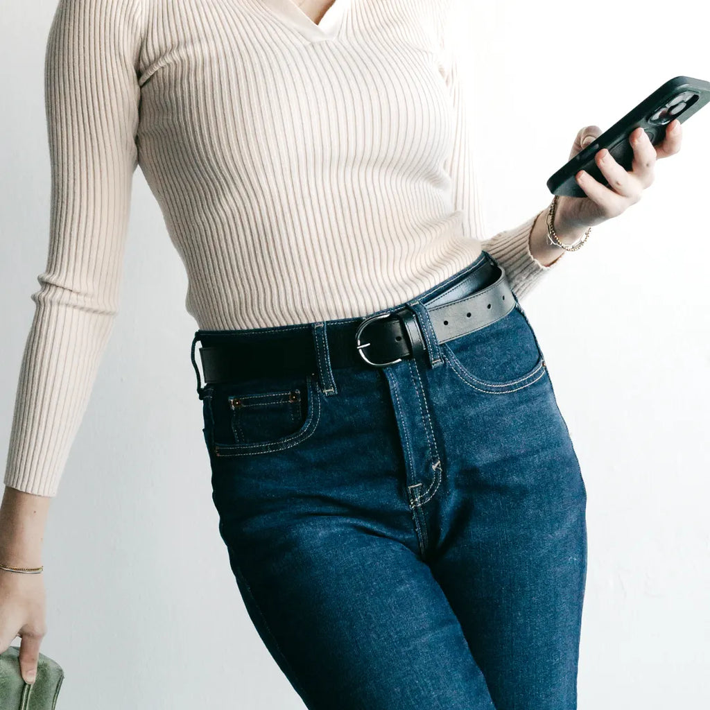 Wearing a cream ribbed long-sleeve top and high-waisted blue jeans styled with the Women’s Matching Leather Belt, this person holds a phone in one hand and a green item in the other.