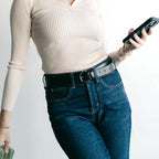 Wearing a cream ribbed long-sleeve top and high-waisted blue jeans styled with the Women’s Matching Leather Belt, this person holds a phone in one hand and a green item in the other.