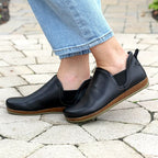A person stands on a stone-paved surface wearing light blue jeans and the Women's Lieben Leather Chelsea Boot—black with brown soles, crafted from vegetable-tanned leather.