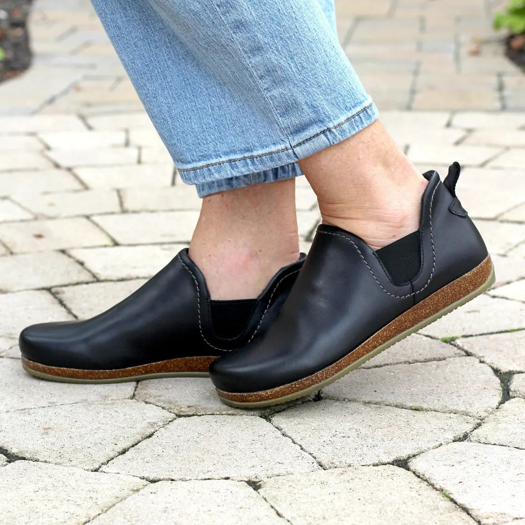 A person stands on a stone-paved surface wearing light blue jeans and the Women's Lieben Leather Chelsea Boot—black with brown soles, crafted from vegetable-tanned leather.