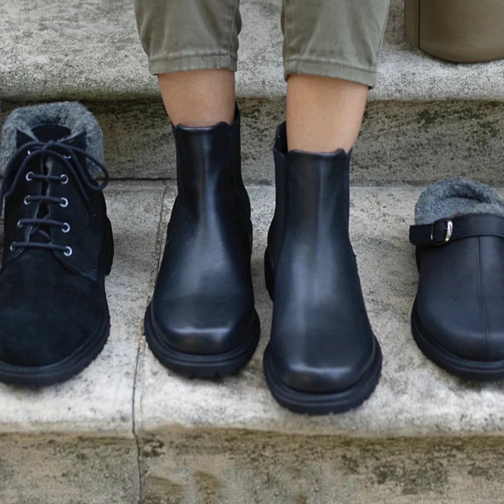 A person wearing khaki pants stands on stone steps, modeling the Women’s Brenner Chelsea Boot—a water-resistant leather ankle boot featuring a sleek black design and gray lining.
