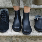 A person wearing khaki pants stands on stone steps, modeling the Women’s Brenner Chelsea Boot—a water-resistant leather ankle boot featuring a sleek black design and gray lining.