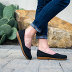 A person in rolled-up jeans stands on a stone path wearing Women’s Liesl Wool Skimmer shoes, featuring a wide toe box and cork soles, with a cactus and large rock in the background.