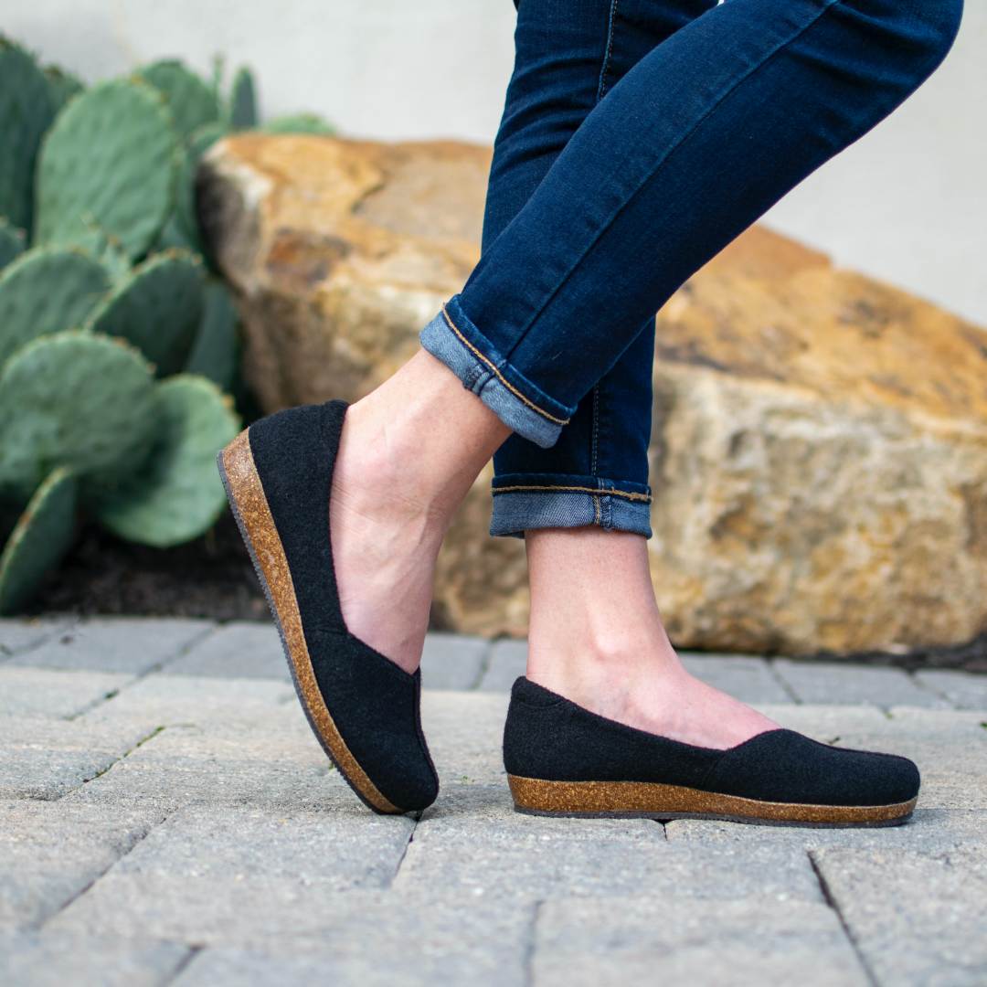 A person in rolled-up jeans stands on a stone path wearing Women’s Liesl Wool Skimmer shoes, featuring a wide toe box and cork soles, with a cactus and large rock in the background.