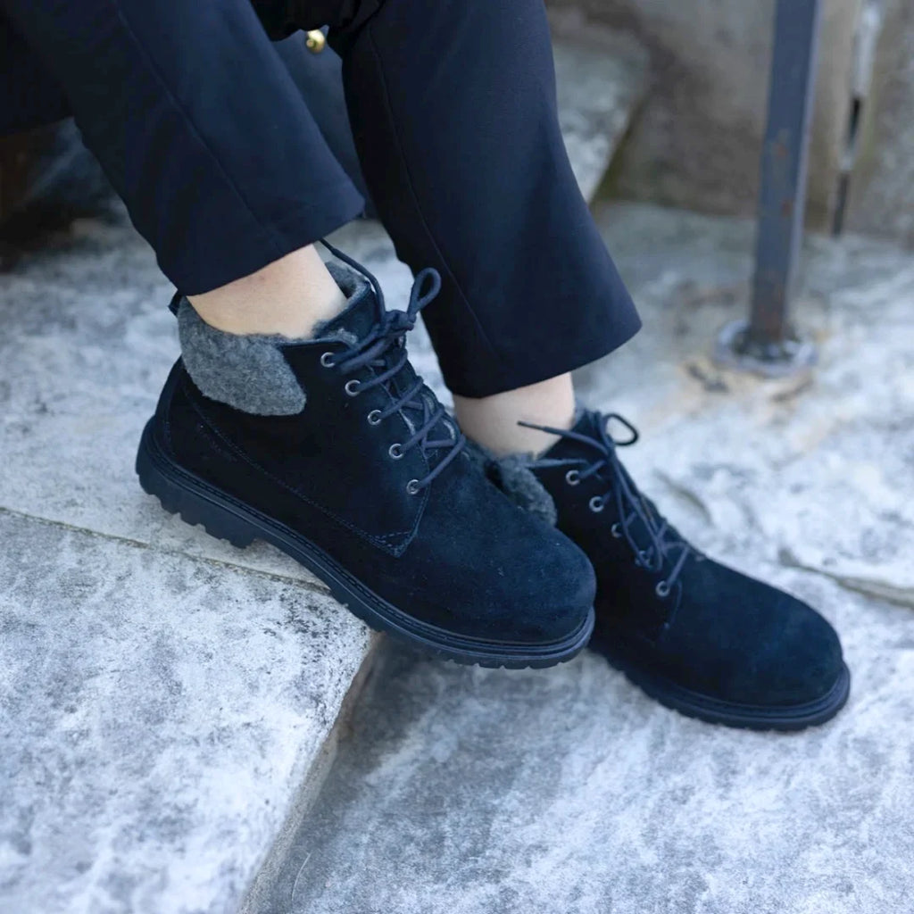 A woman wearing the Women’s Merano Chukka Boot—black lace-up boots with grey fleece lining—sits on concrete steps, with only her lower legs and feet visible.