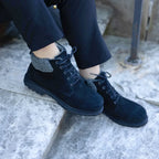 A woman wearing the Women’s Merano Chukka Boot—black lace-up boots with grey fleece lining—sits on concrete steps, with only her lower legs and feet visible.