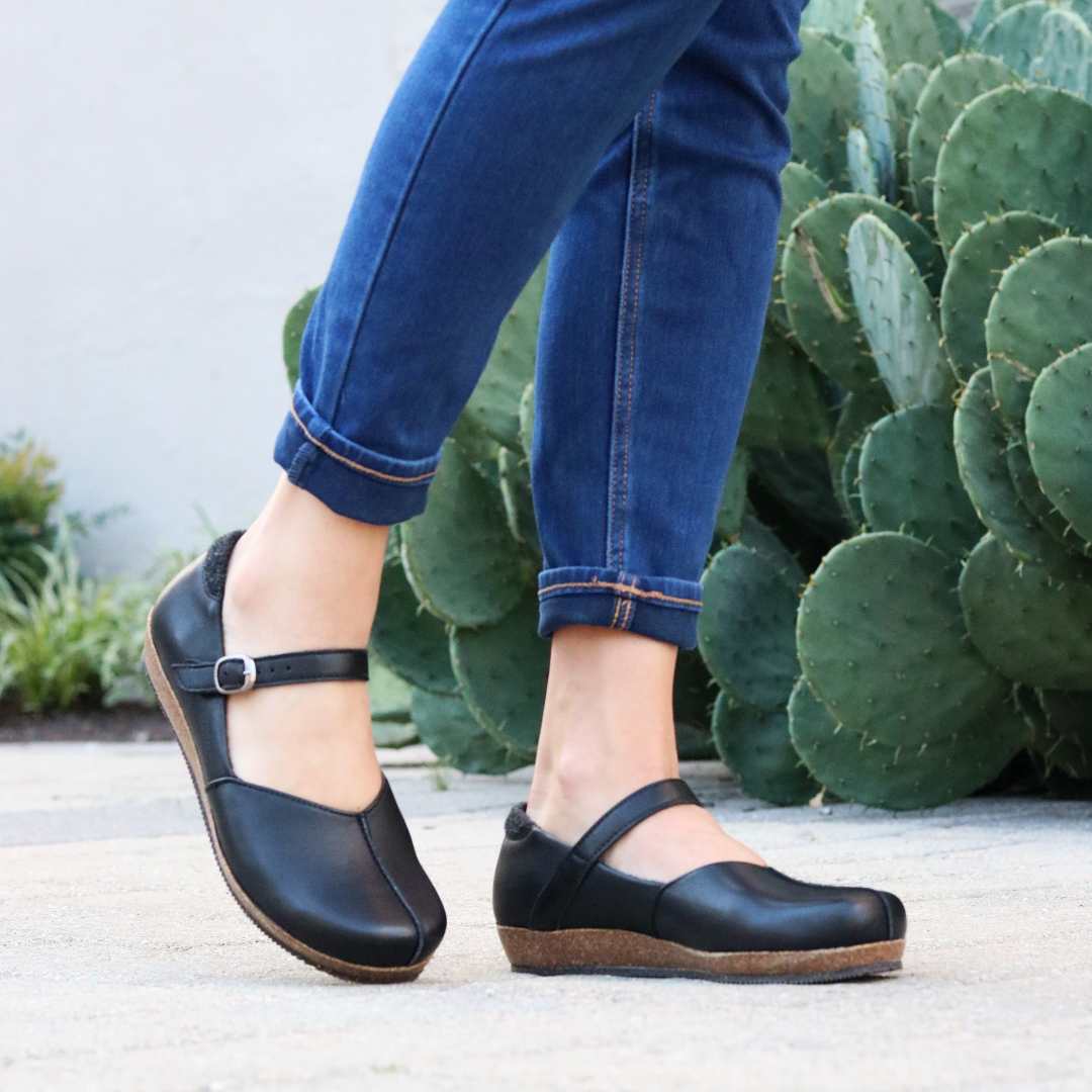 A person wearing Women's Eva Leather Mary-Jane shoes with cork soles and a wide toe box, paired with blue jeans, stands on a paved surface beside a large cactus.
