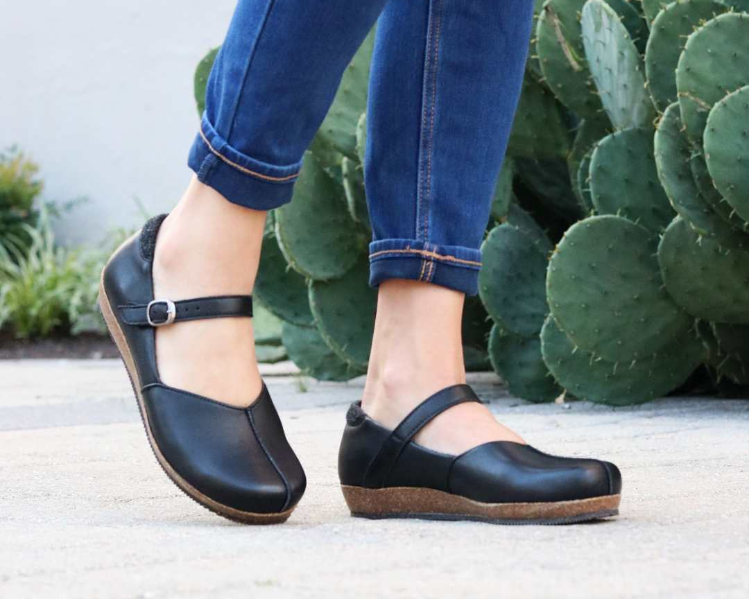 A person stands on a paved surface near a large cactus, wearing cuffed blue jeans and Women's Eva Leather Mary-Jane shoes with wide toe boxes and cork soles.