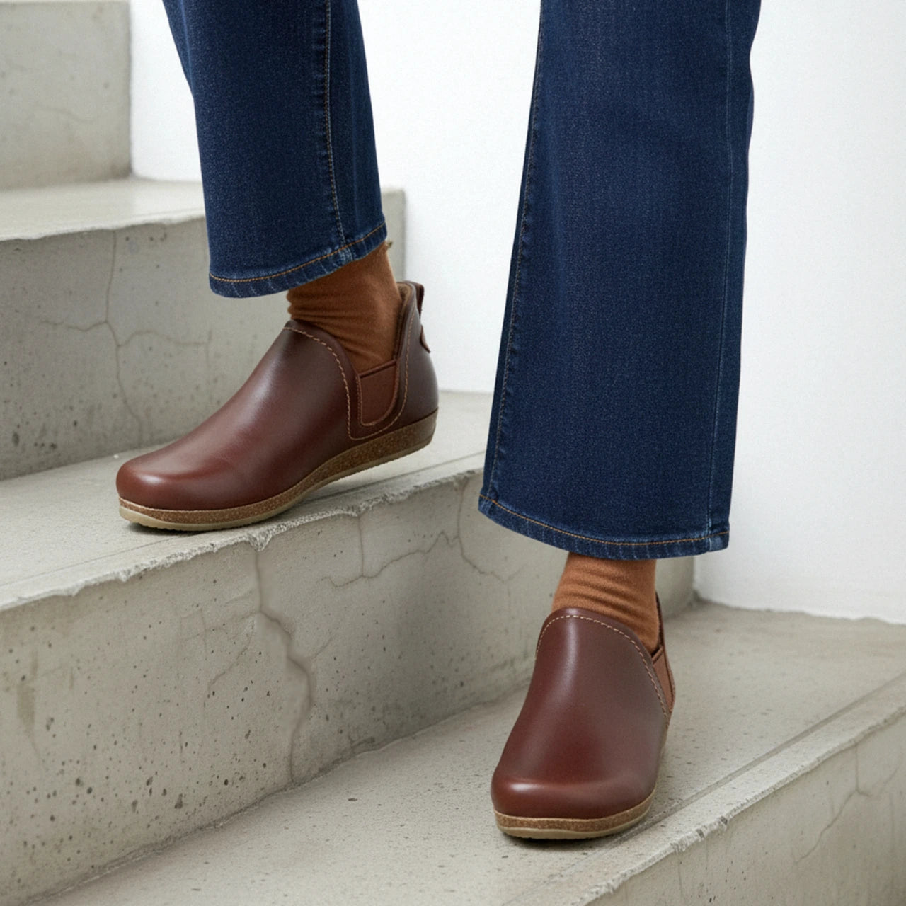 A person wearing the Women's Lieben Leather Chelsea Boot with brown socks and blue jeans stands on concrete stairs.