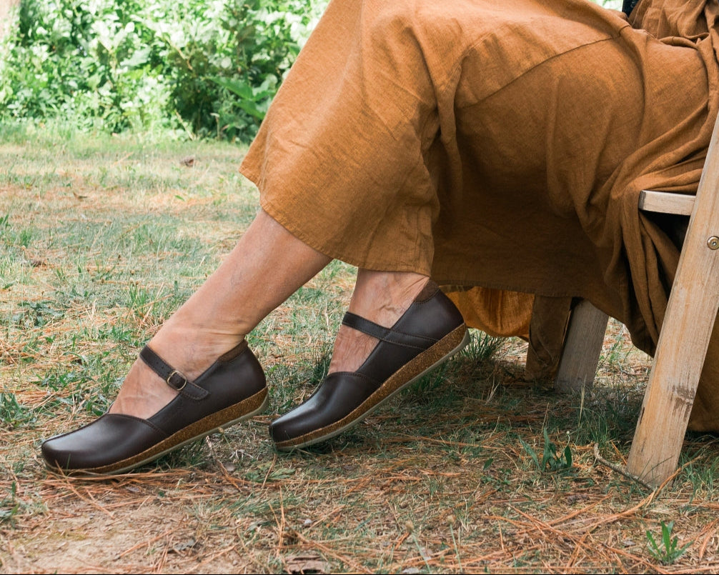 A person sits outdoors on a wooden chair, wearing a long brown skirt and Women's Eva Leather Mary-Jane shoes, surrounded by greenery.