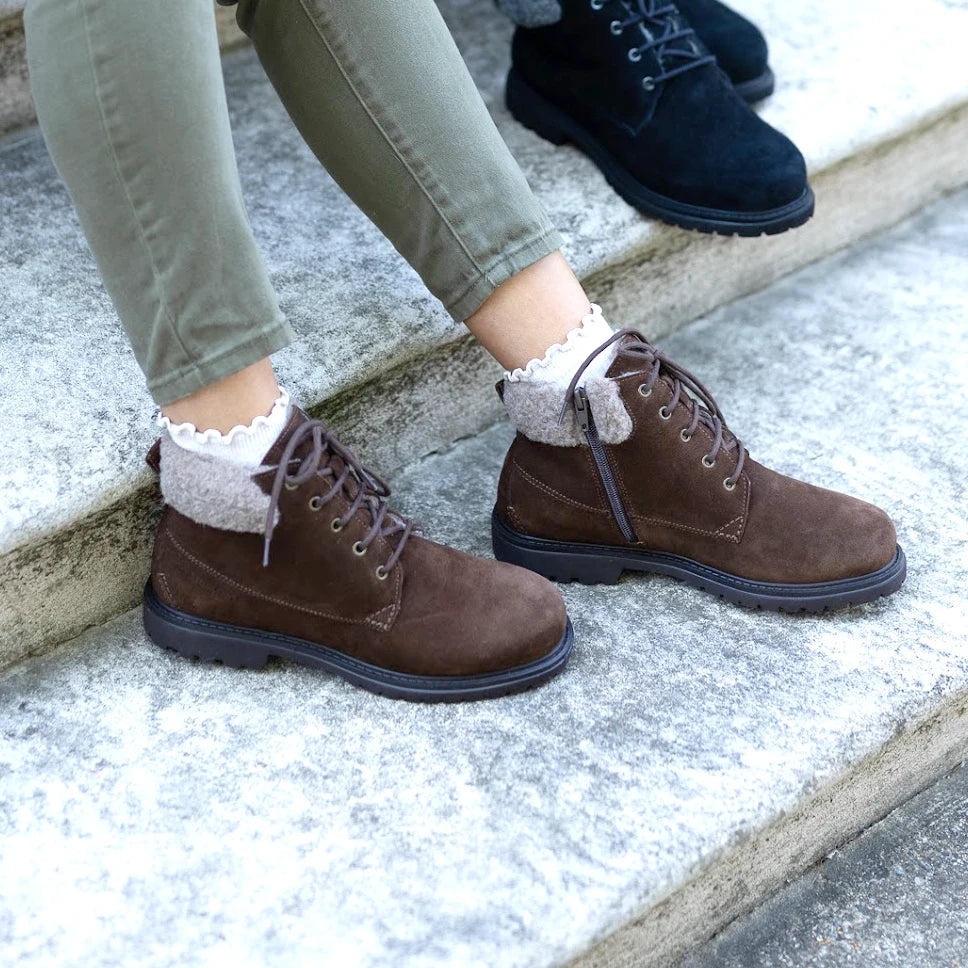 A person wearing the Women’s Merano Chukka Boot in brown waterproof suede with light-colored socks and olive green pants sits on outdoor stone steps, while another individual in black boots is partially visible in the background.