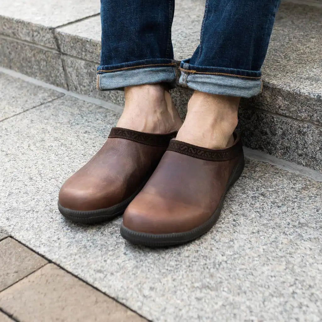 A person wearing rolled-up jeans and brown Men's Graz Leather Clog with Sport Sole, featuring wool felt lining, sits on stone steps.