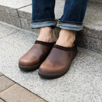 A person wearing rolled-up jeans and brown Men's Graz Leather Clog with Sport Sole, featuring wool felt lining, sits on stone steps.