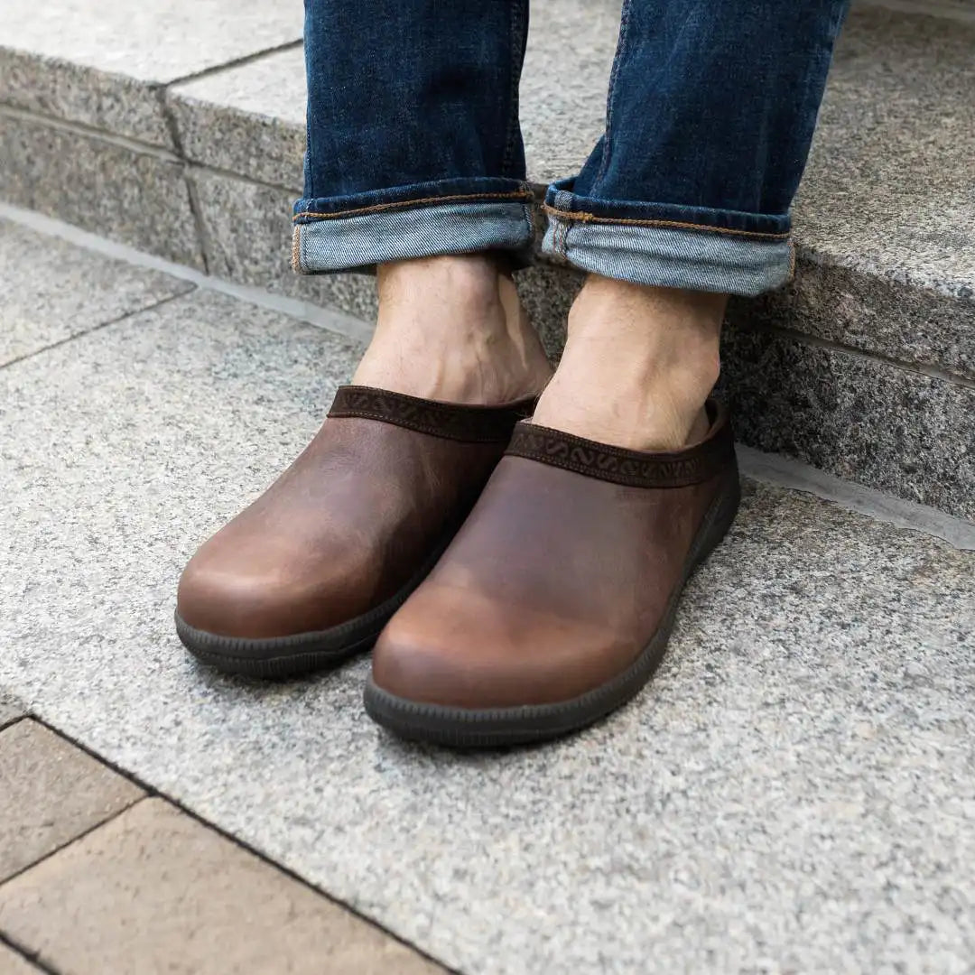 A person wearing rolled-up jeans and brown Men's Graz Leather Clog with Sport Sole, featuring wool felt lining, sits on stone steps.